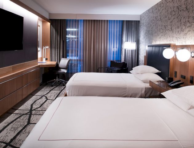The image shows a modern hotel room with two twin beds, a flat-screen TV, a work desk, and large windows with curtains.