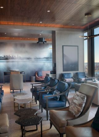 The image features a modern lounge area with leather chairs, a mural of a cityscape, and large windows offering a scenic view of a waterfront.