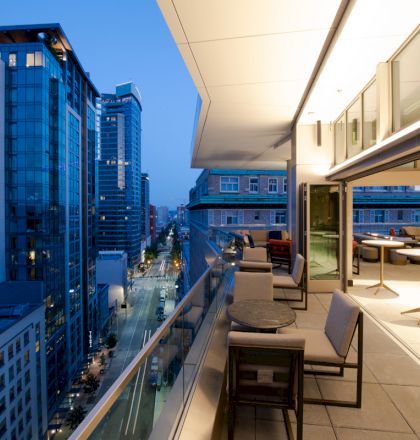 This image shows a modern cityscape with high-rise buildings and a rooftop terrace featuring tables and chairs, overlooking a street at dusk.