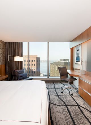This image shows a modern hotel room with a large bed, wall-mounted TV, desk, chair, and large windows offering a city and water view.