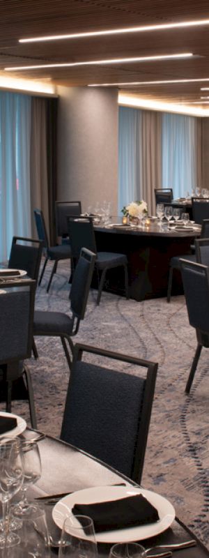 A banquet hall is set up with round tables covered in dark tablecloths, chairs, glassware, and flower centerpieces, under a ceiling with modern lighting.