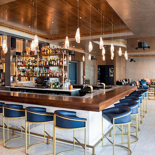 A modern bar with a well-stocked liquor shelf, blue bar stools, pendant lights, and a spacious, elegantly designed interior.