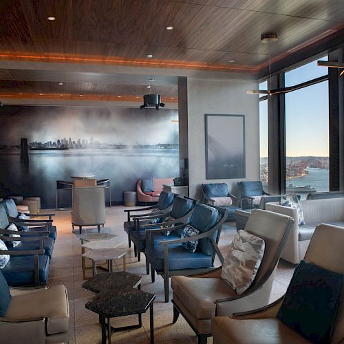 A stylish lounge area with multiple chairs, tables, a wall mural, and large windows offering a scenic view of the waterfront and Ferris wheel.