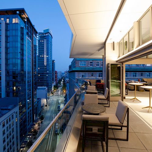 A modern urban balcony overlooks a cityscape at dusk, featuring comfortable seating and tables, with buildings and a street below.