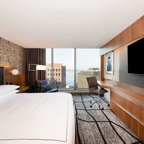 This is a modern hotel room featuring a bed, large wall-mounted TV, desk, chair, and large window with a city view.