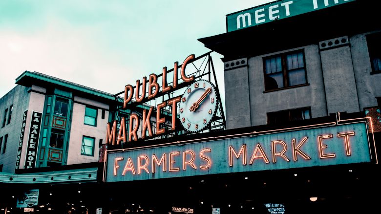 The image shows a neon sign reading "Public Market" and "Farmers Market" on a building. Another sign reads "Meet the...(rest obscured)."
