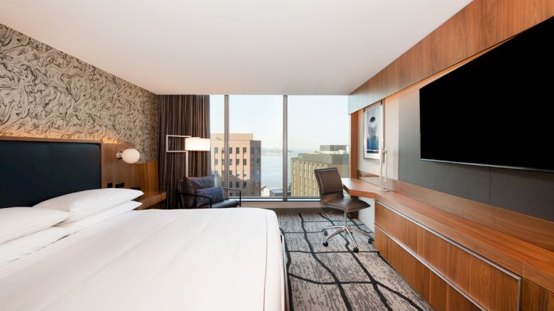 A modern hotel room with a large bed, wall-mounted TV, desk, and chair, featuring a city view through floor-to-ceiling windows.