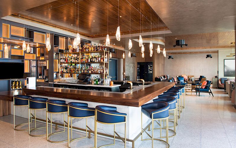 A modern bar with a well-stocked shelf, surrounded by blue and gold bar stools, hanging lights, and a lounge area with seating in the background.