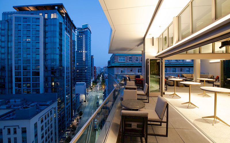A modern balcony overlooks a cityscape with tall buildings, featuring comfortable seating and tables, while the evening sky adds to the ambience.
