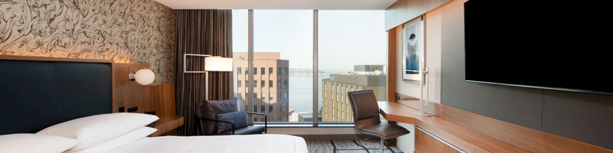 A modern hotel room with a neatly made bed, wall-mounted TV, desk, and chair, featuring large windows with a city view in the background.
